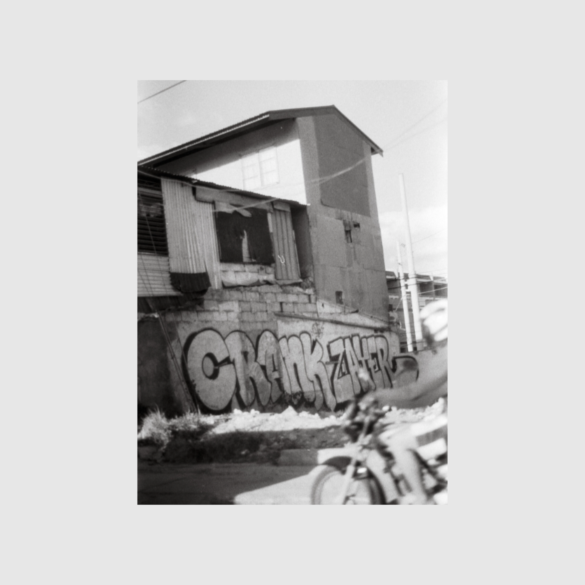 A black and white photo of a rundown building with graffiti reading "CRANKZAYER" on the wall. A person on a motorcycle is blurred in the foreground.