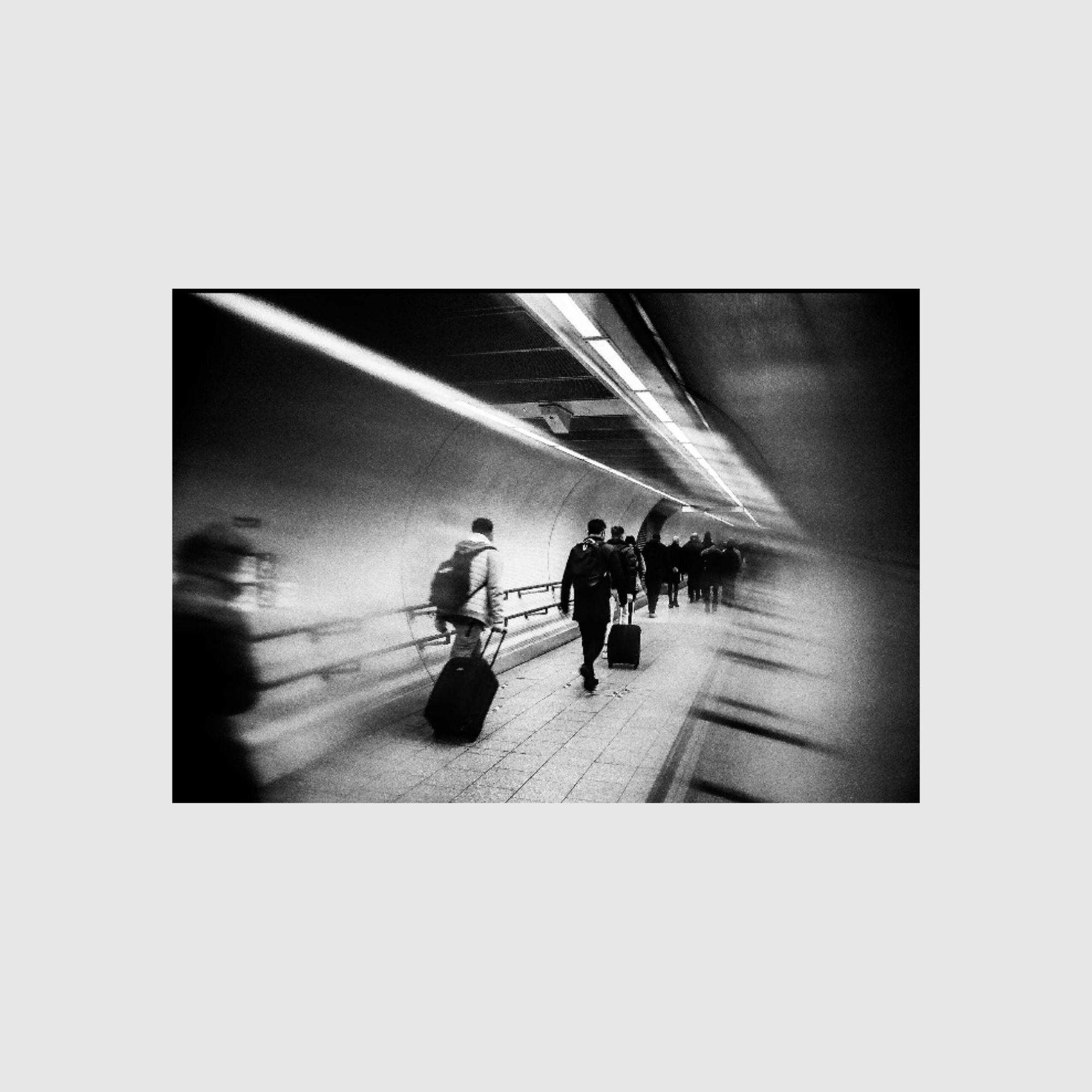 Black and white photo of people walking through a tunnel, with some carrying luggage. The scene captures motion and blur, giving a sense of hurried movement and travel. The tunnel is illuminated by overhead lights, creating a dynamic, almost surreal atmosphere.