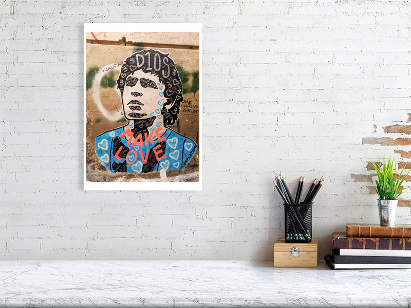 A framed art print of Diego Maradona street art featuring the inscription 'D10S' and the phrase 'MAKE LOVE' displayed on a white brick wall above a desk with a pencil holder, books, and a small plant