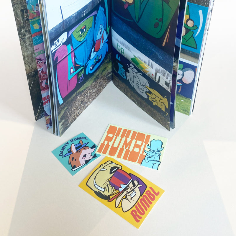 Open pages of the book/zine 'Hanna Barbarian II' by Danny Rumbl, displaying colorful illustrations of cartoon characters painted as murals on outdoor surfaces. In front of the book are three colorful stickers featuring cartoon characters and the name 'Rumbl' in bold letters.