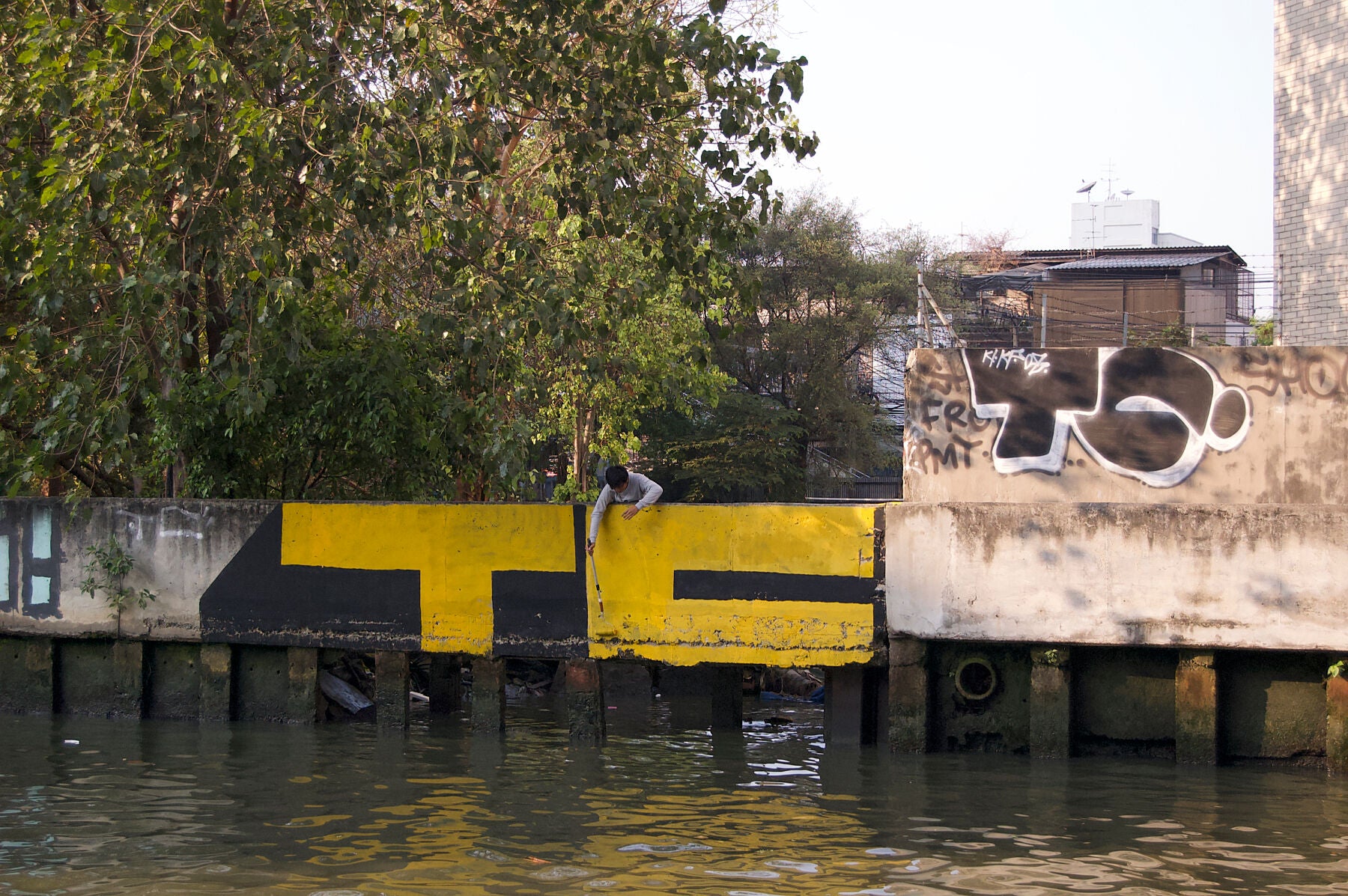 A man from TC crew painting a large yellow and black graffiti mural that reads TC, on a concrete wall by the water, with trees and buildings in the background.