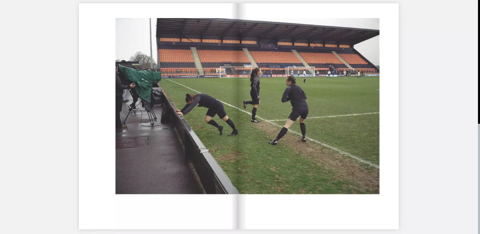 Two-page spread from the zine "The Women's Game" by Claire Wray. The image shows three female football players in black training gear stretching and warming up on the sideline of a football field. In the background, there is a nearly empty stadium with orange and black seats. On the left side of the image, a camera crew is filming, with one cameraman adjusting the equipment under a green cover. The scene appears to be during a practice or pre-match preparation.