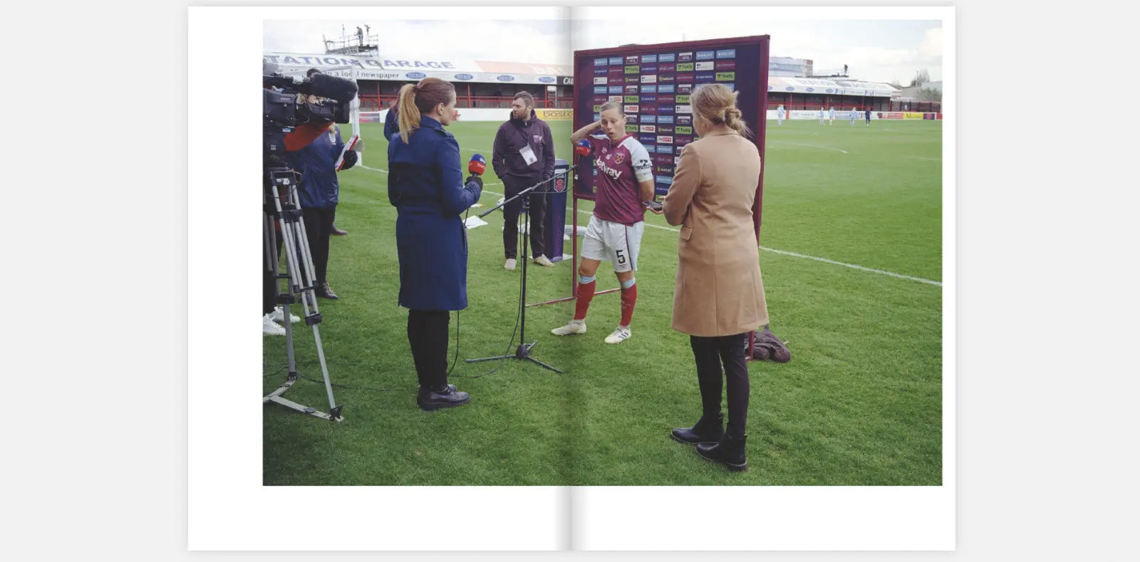 Two-page spread from the zine "Women's Football" by Claire Wray. The image shows a post-match interview on a football field. A female football player in a maroon and white uniform stands in front of a media backdrop, speaking into microphones held by two female reporters. One reporter wears a blue coat, and the other wears a tan coat. A cameraman films the scene while another person looks on. The setting is a stadium with green grass and stands in the background.